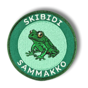 Brodeerattu haalarimerkki skibidi sammakosta saumatulla reunalla, tekstillä: ''Skibidi sammakko''.